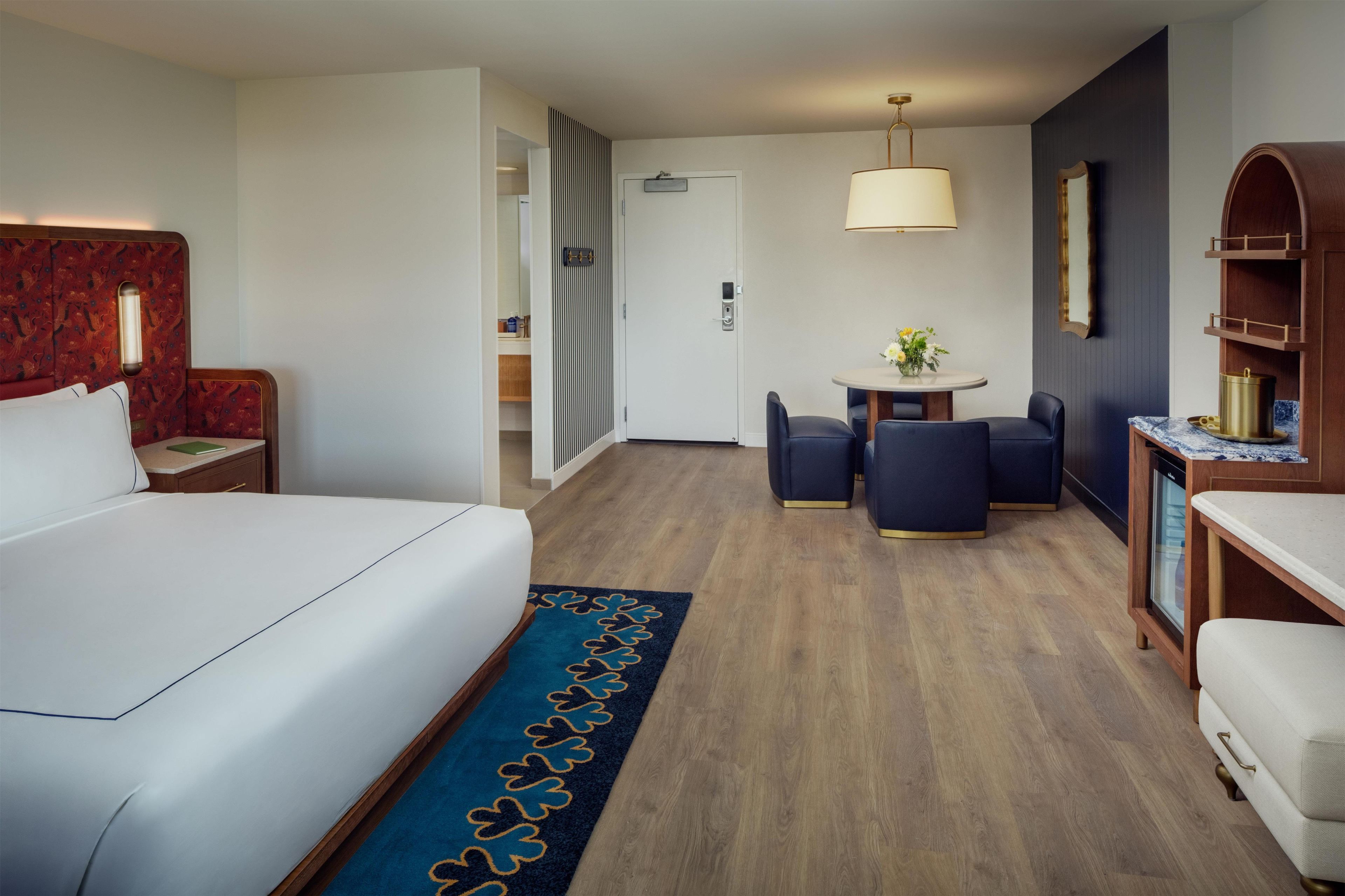 A hotel room with a large bed, a desk with two chairs, a small table with a lamp, and a wooden cabinet.