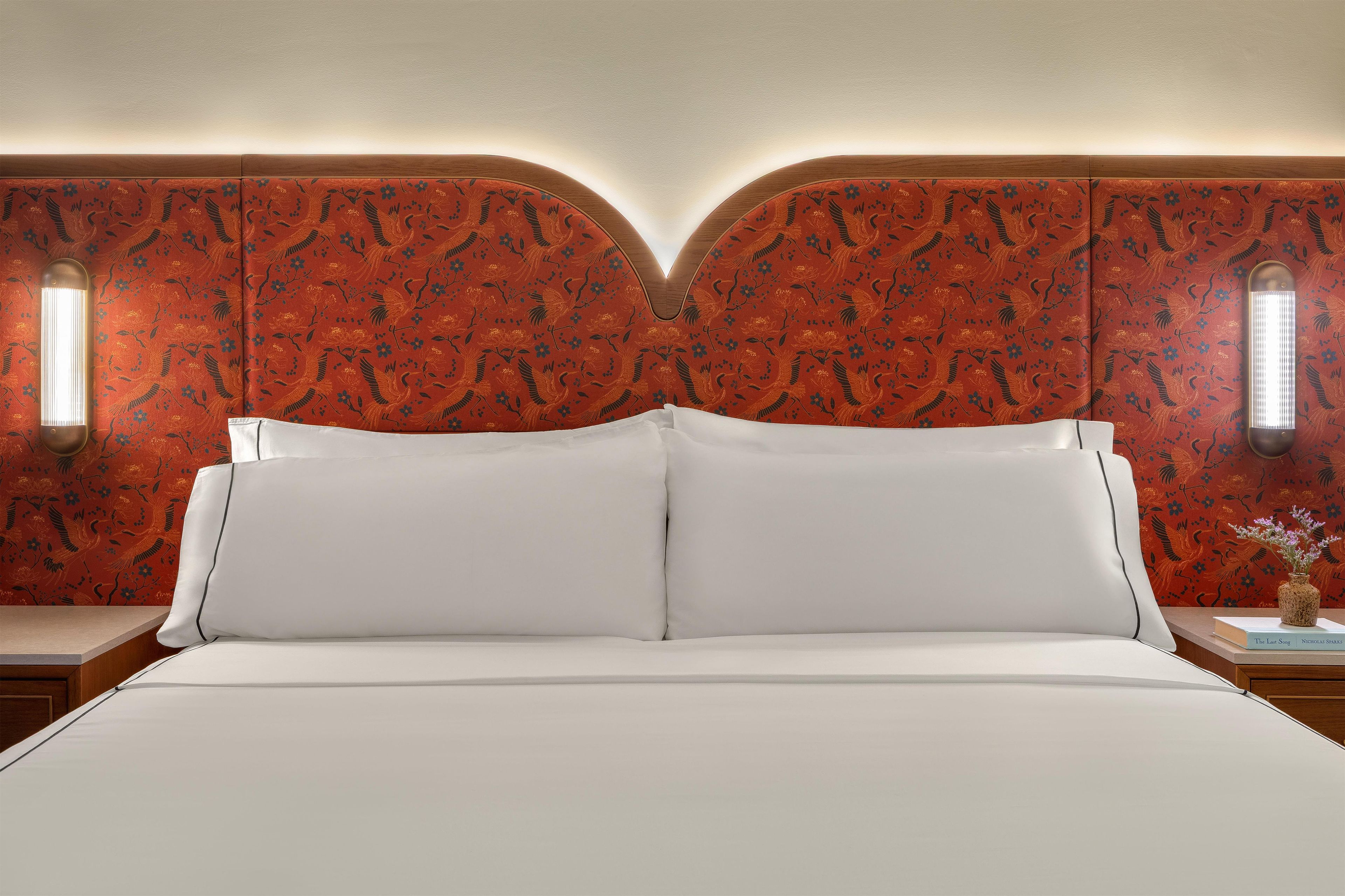 Ein Bett mit weißen Laken in einem Raum, der ein rot gemustertes Kopfteil und zwei Wandleuchten hat.