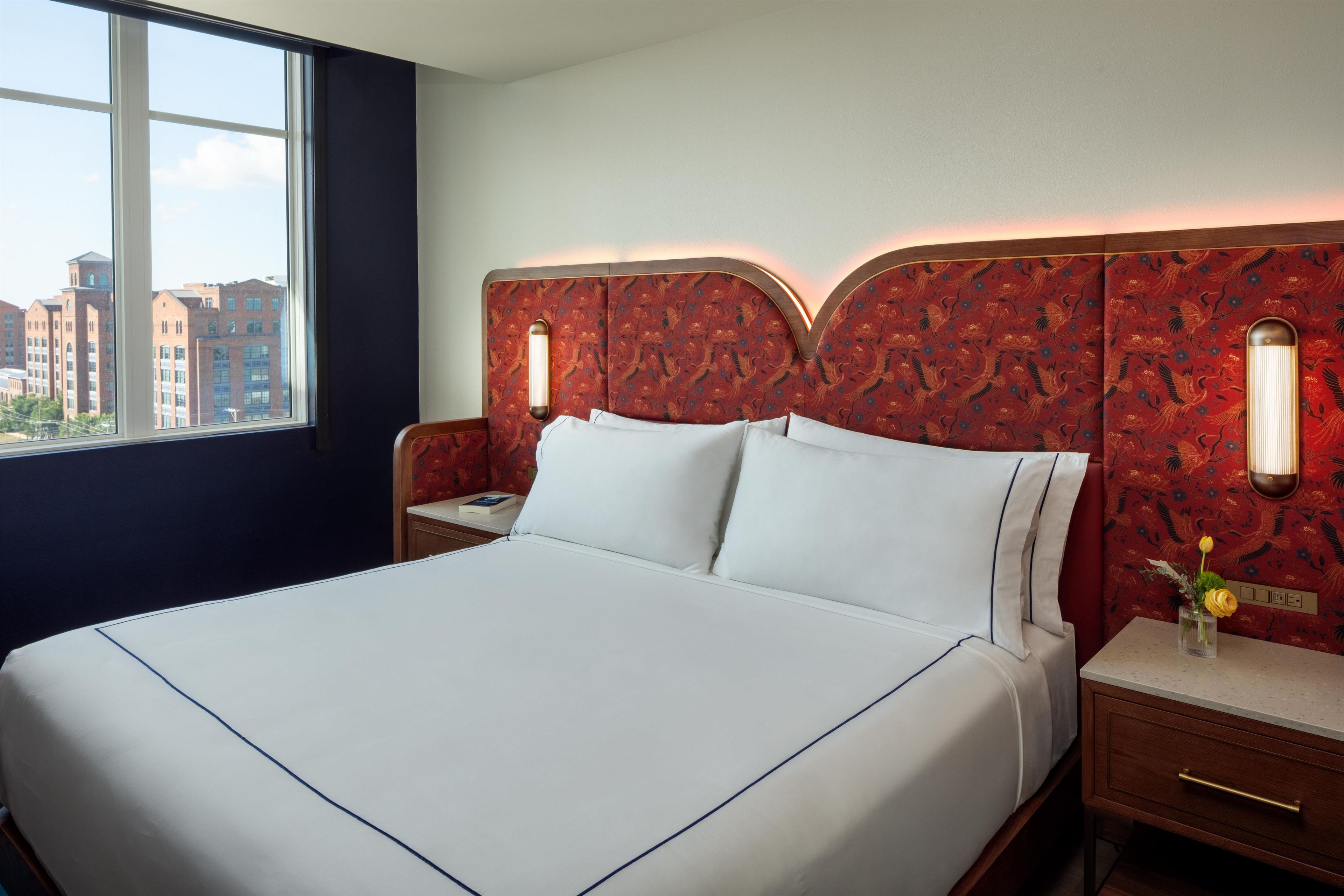 Une chambre d’hôtel avec un grand lit, une table de chevet, une commode, une fenêtre donnant sur la ville et une tête de lit à motifs.