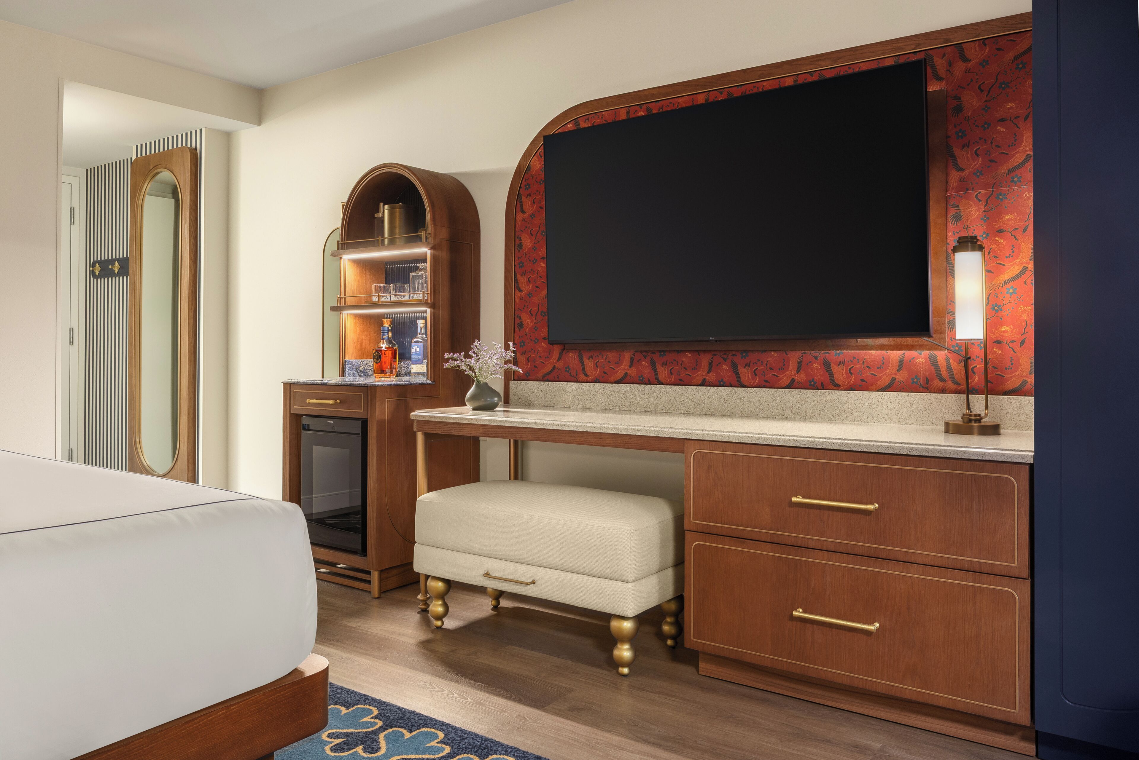 Habitación de hotel con cama, escritorio con cajones, televisor, espejo y repisa con botellas.
