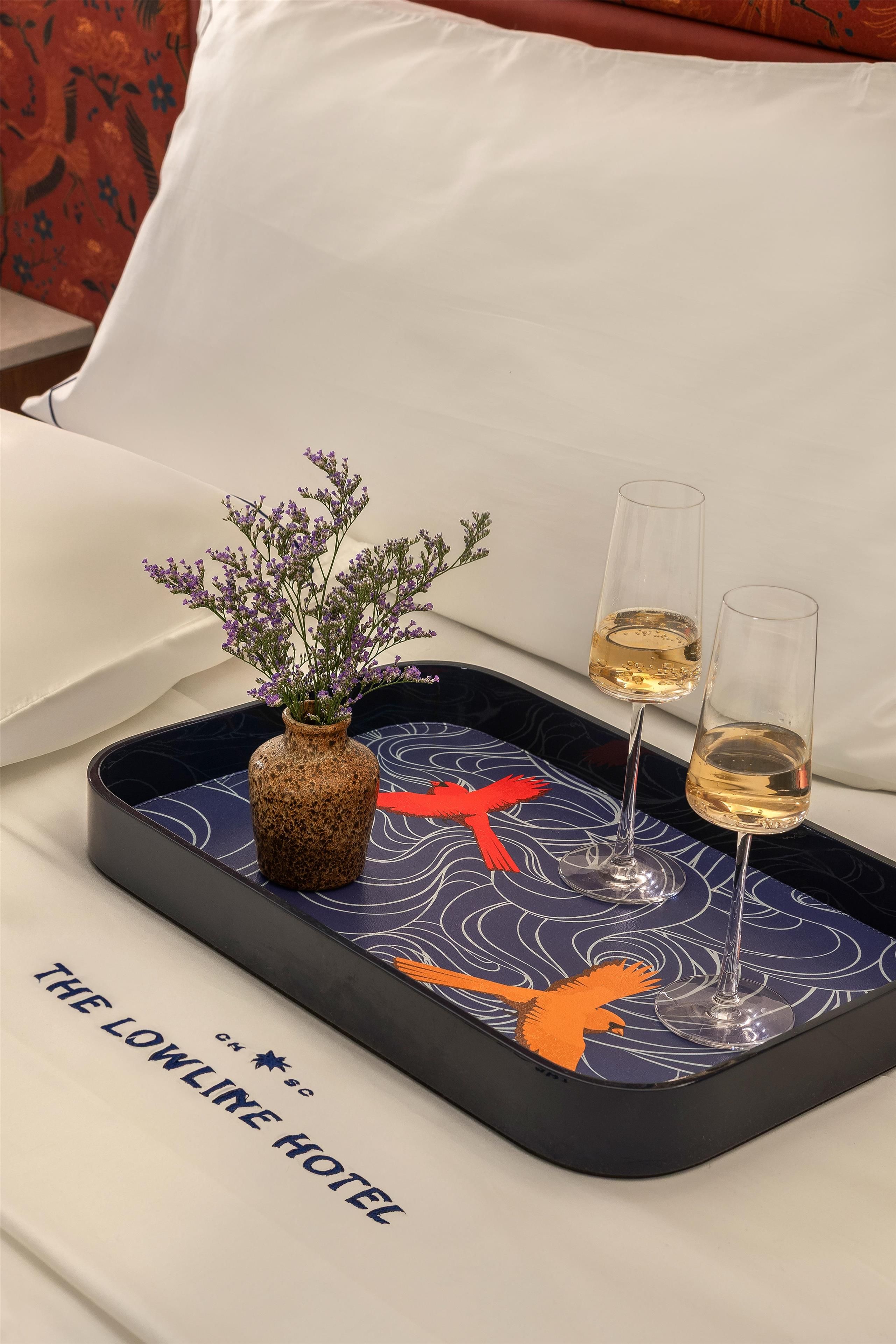 A tray with a vase of flowers and two glasses of wine on a bed with a pillow and a decorative pillowcase.
