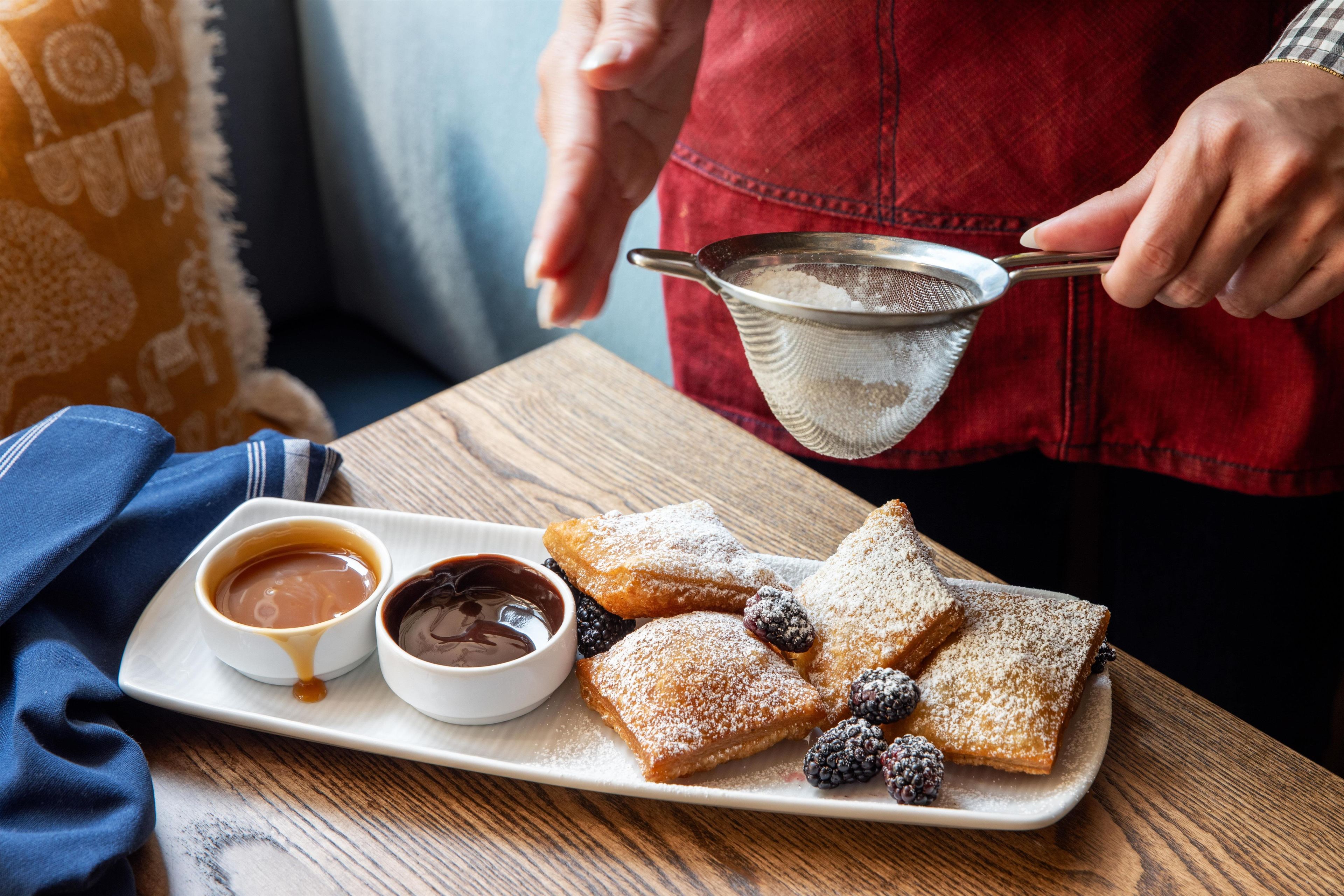 A person holding a strainer over a plate of pastries with powdered sugar, accompanied by two small bowls of dipping sauces.