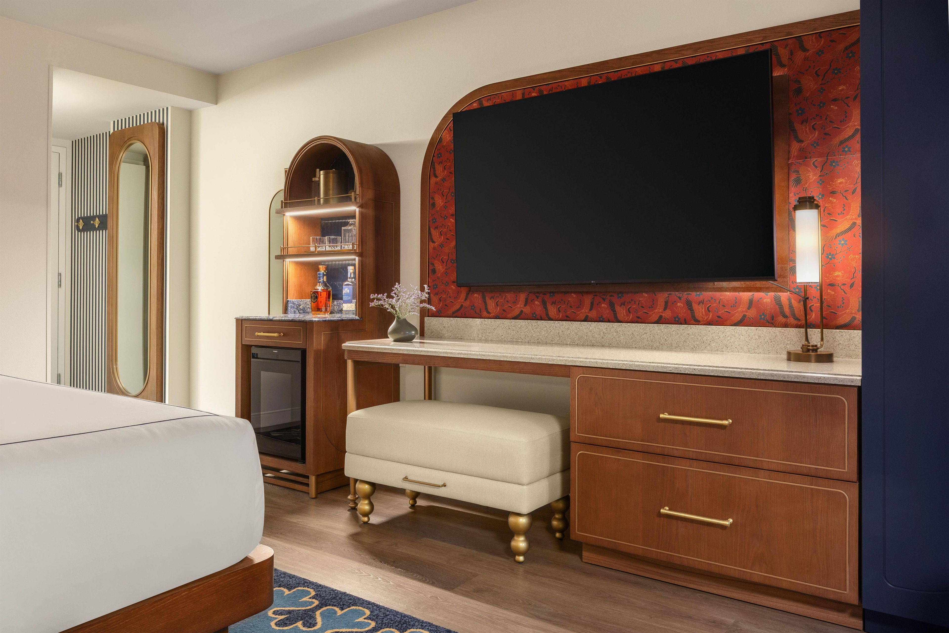 A hotel room with a bed, a desk with a flat-screen TV, a vanity with a mirror, and a small shelf with toiletries.