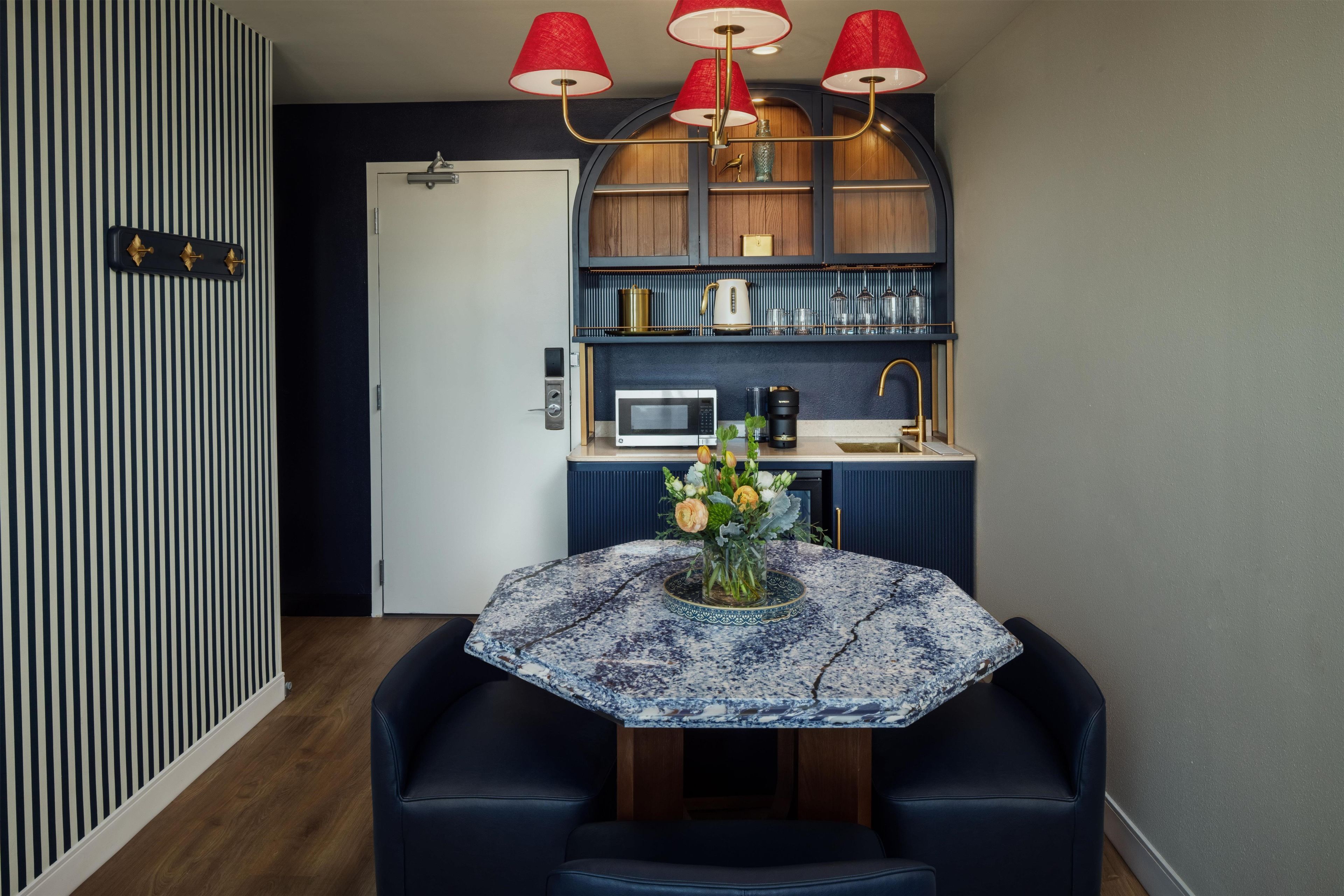 Una cocina compacta con microondas, cafetera y un lavamanos pequeño. Hay una mesa redonda con un florero y dos sillas.