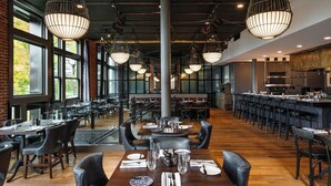 A spacious restaurant with high ceilings, exposed brick walls, and large windows. Tables are set with white tablecloths and black chairs. - The Charmant Hotel (La Crosse)