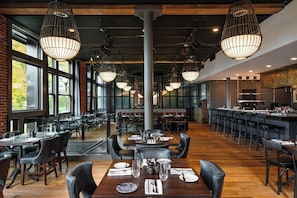 A spacious restaurant with high ceilings, exposed brick walls, and large windows. Tables are set with white tablecloths and black chairs. - The Charmant Hotel (La Crosse)