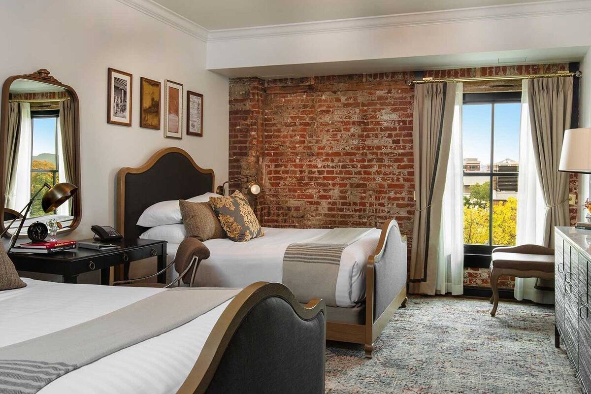 A hotel room with two beds, a brick wall, a large window with curtains, and a desk with a lamp.
