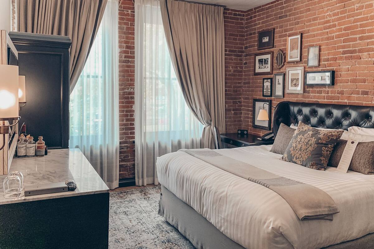 A hotel room with a brick wall, a bed with multiple pillows, a desk with a lamp, and a window with curtains.