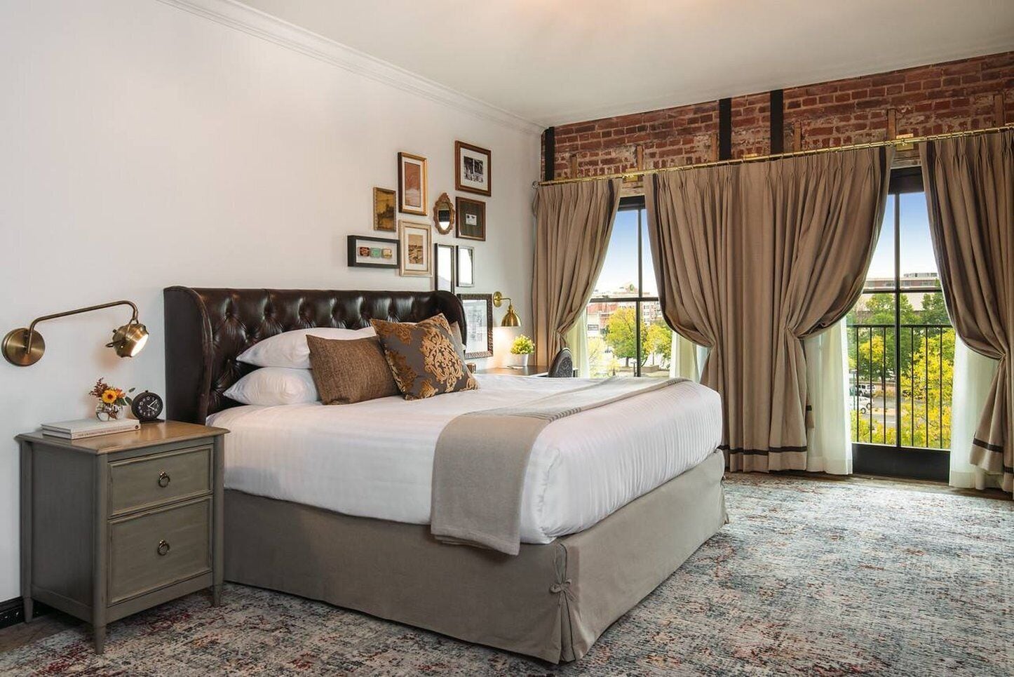 A bedroom with a large bed, a nightstand, a wall-mounted lamp, and a view through large windows with curtains.