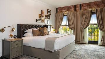 A bedroom with a large bed, a nightstand, a wall-mounted lamp, and a view through large windows with curtains.