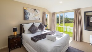 A bedroom with a large bed, a nightstand, a lamp, and a view of a green landscape through the window. - The Meadows Villa (Christchurch)