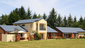 A modern two-story house with a flat roof, a patio with outdoor furniture, and a well-maintained lawn. - The Meadows Villa (Christchurch)