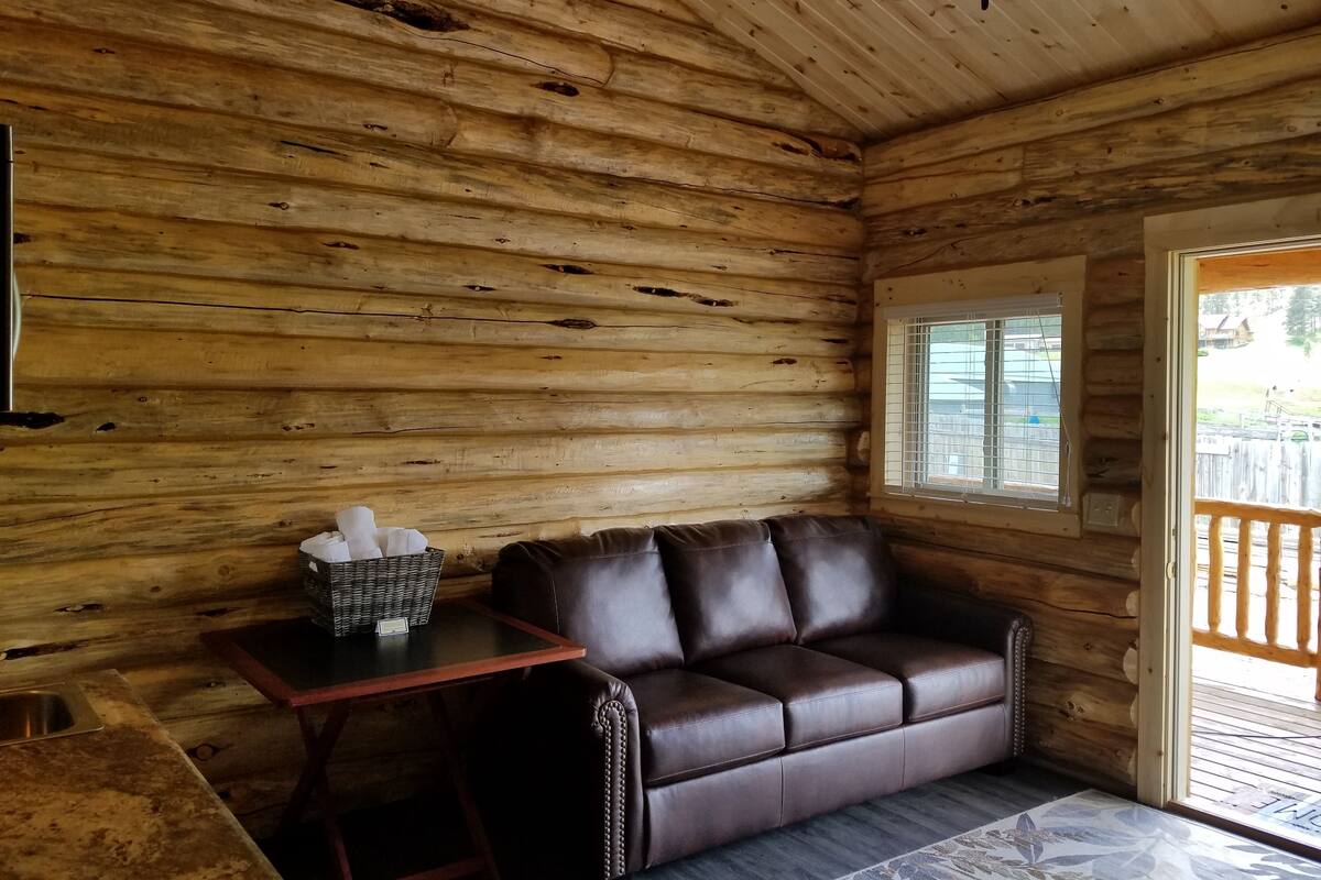 A log cabin interior with a leather sofa, a small table with a basket, and a window with a view of the outside.