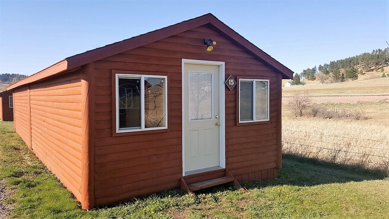 A small, single-story wooden cabin with a white door and two windows, set in a grassy field with a clear sky.