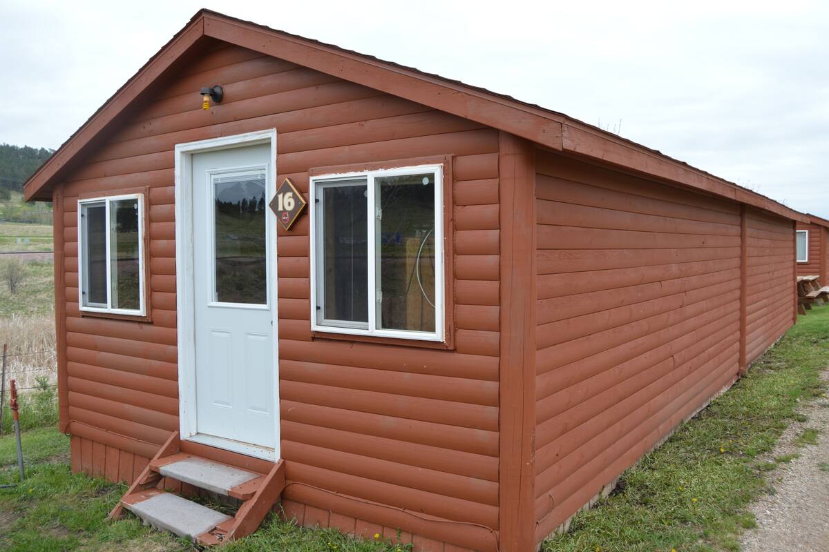 A small, single-story wooden cabin with a white door and two windows, a small porch, and a number '16' sign on the door.