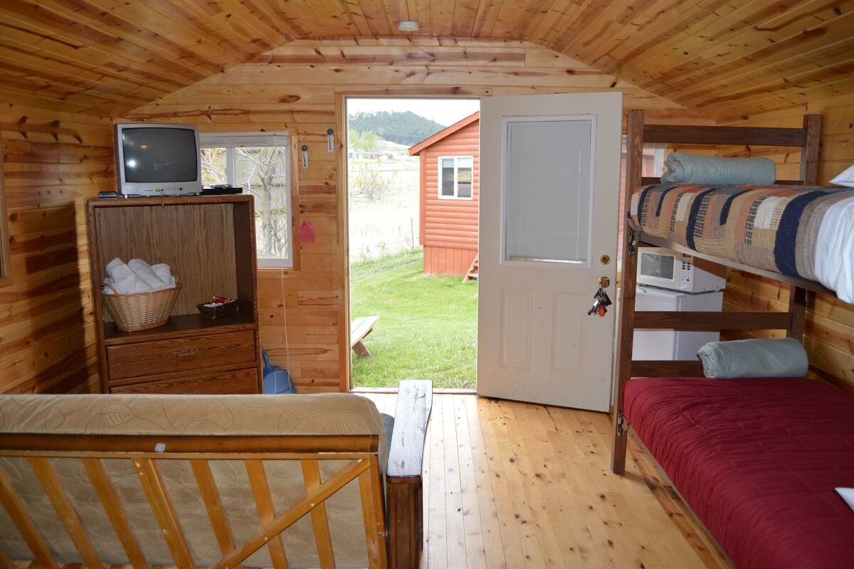 A wooden cabin with a bunk bed, a desk with a television, and a door leading to a grassy yard.