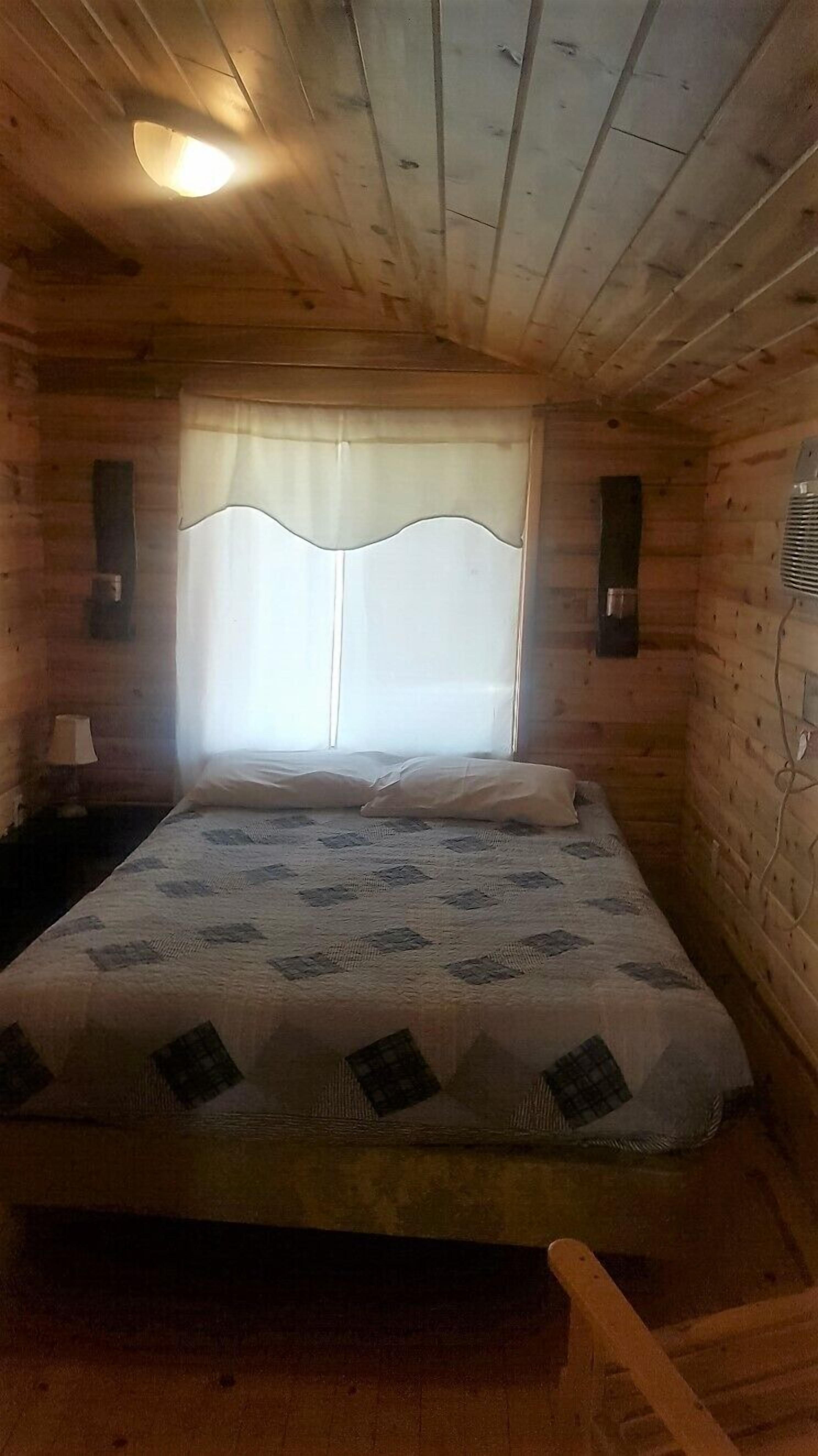 A wooden cabin room with a bed, a window with curtains, a nightstand, and a wall-mounted heater.
