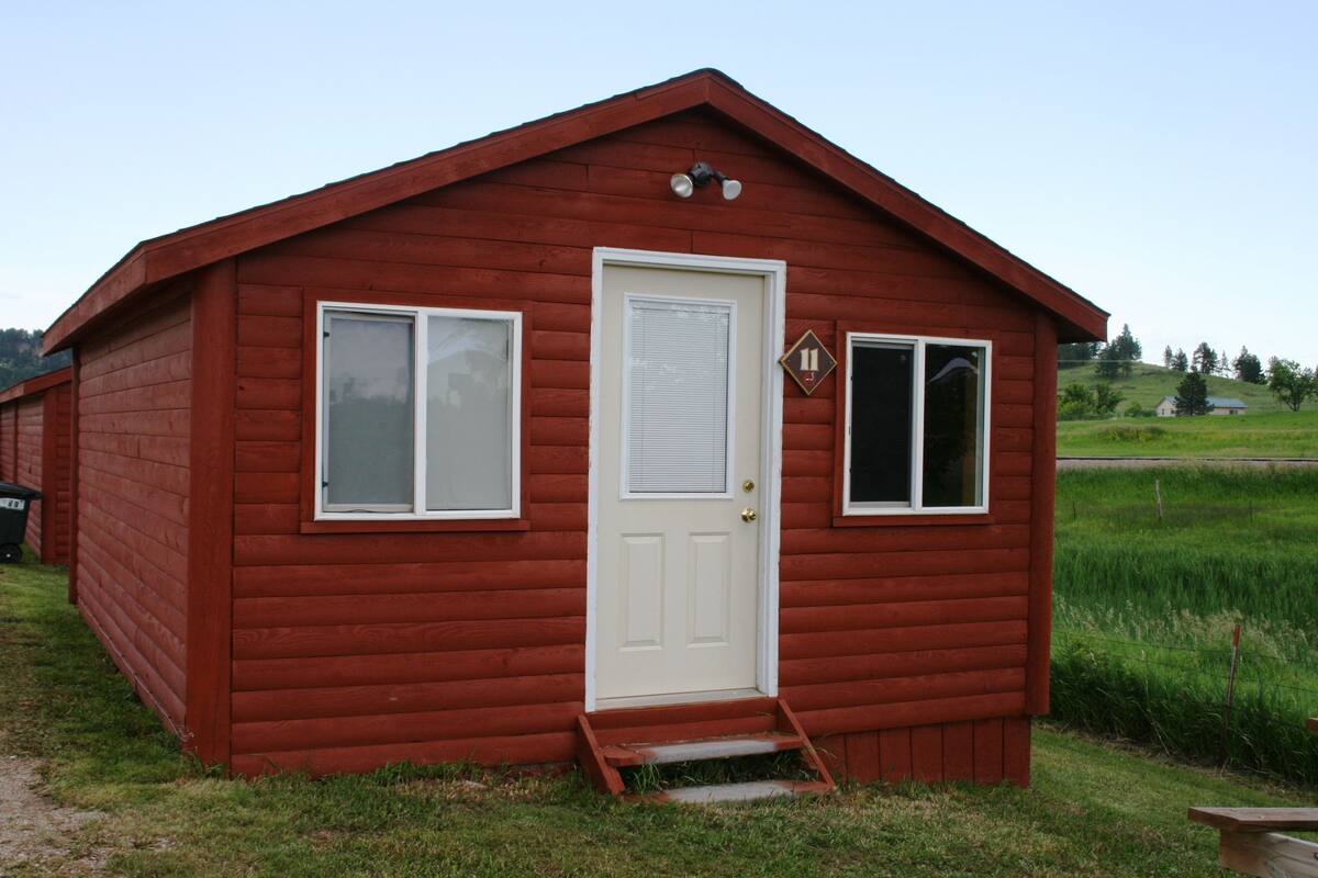 A small, red wooden cabin with a white door, two windows, and a small porch.