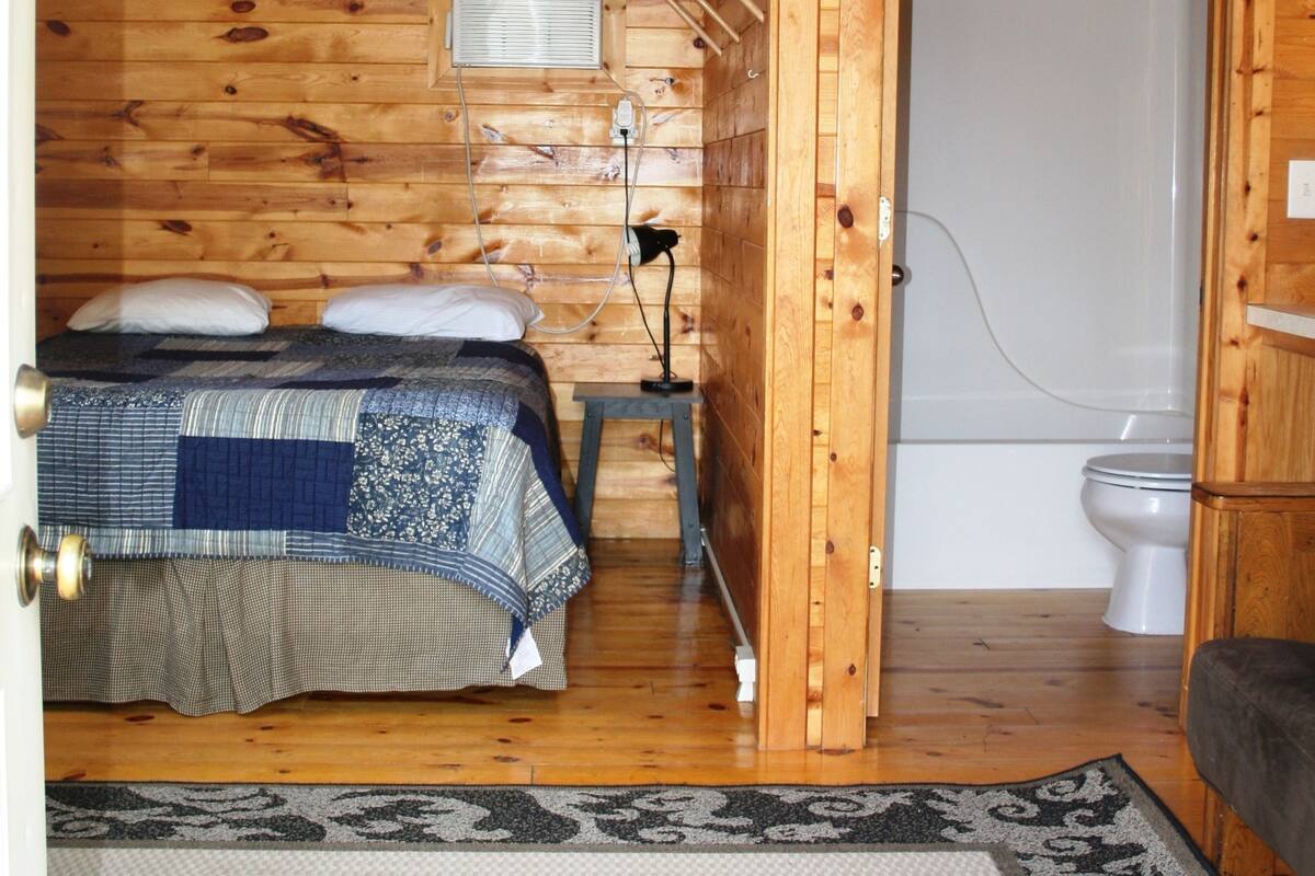 A wooden cabin room with a bed, a small table, a heater, and a bathroom.