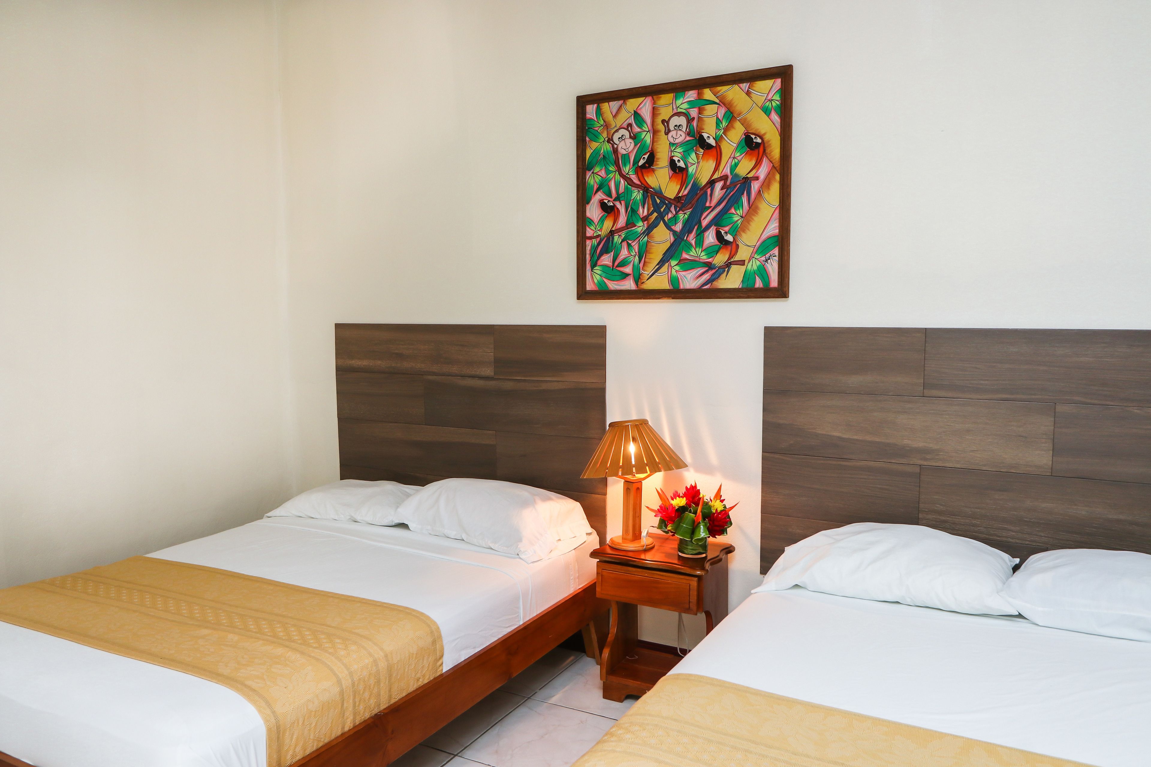Habitación de hotel con dos camas individuales, una mesita de noche con lámpara y flores, y un cuadro colorido en la pared.