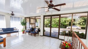 A spacious living area with a blue sofa, wooden chairs, a coffee table, and a ceiling fan. Large windows offer a view of the street and parked cars. - Hotel Flor Blanca (Quepos)