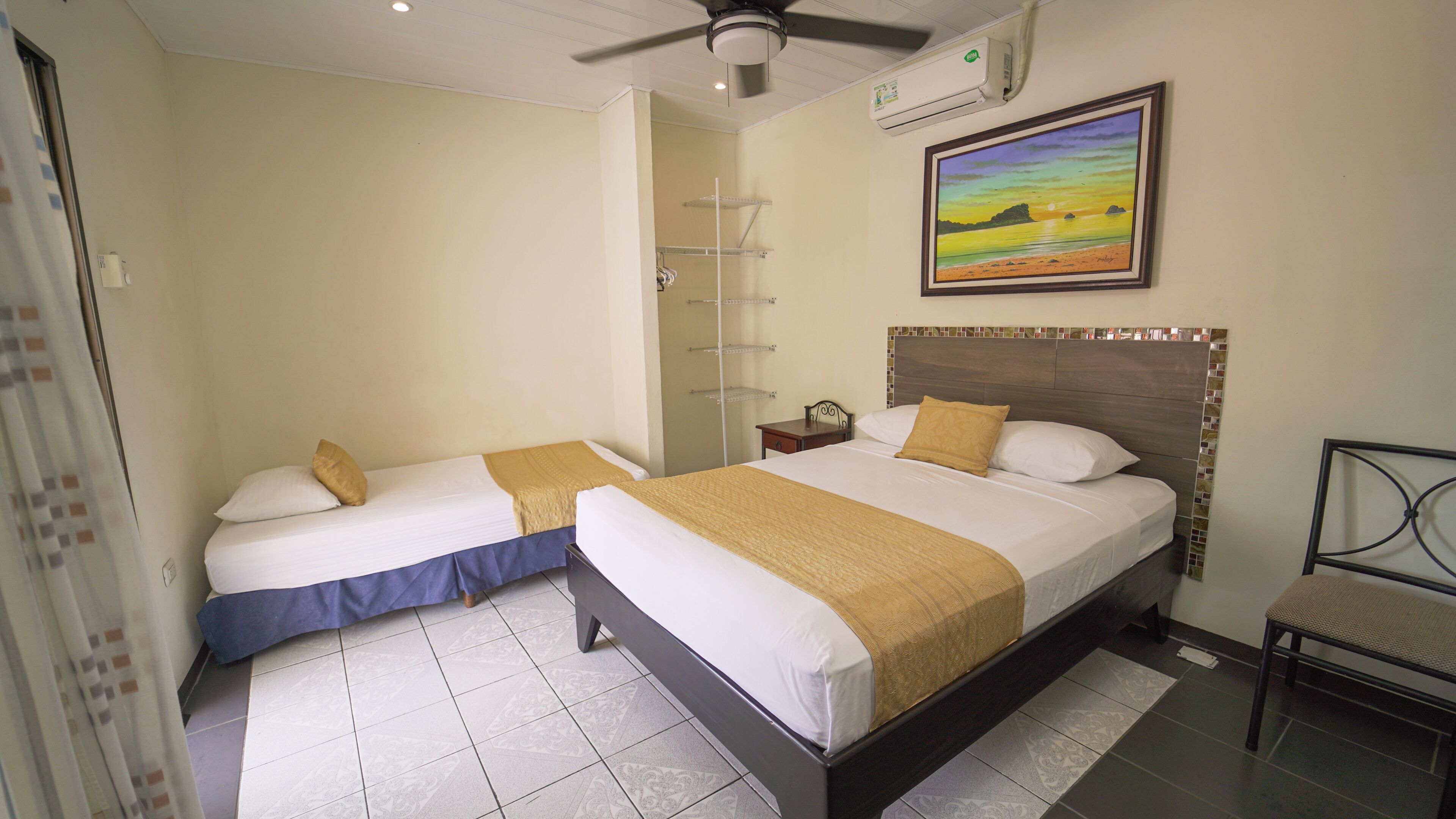 A hotel room with two beds, a ceiling fan, an air conditioning unit, a painting, and a tiled floor.