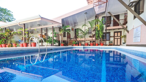 A swimming pool with a tiled floor, surrounded by potted plants and a building with balconies. - Hotel Flor Blanca (Quepos)