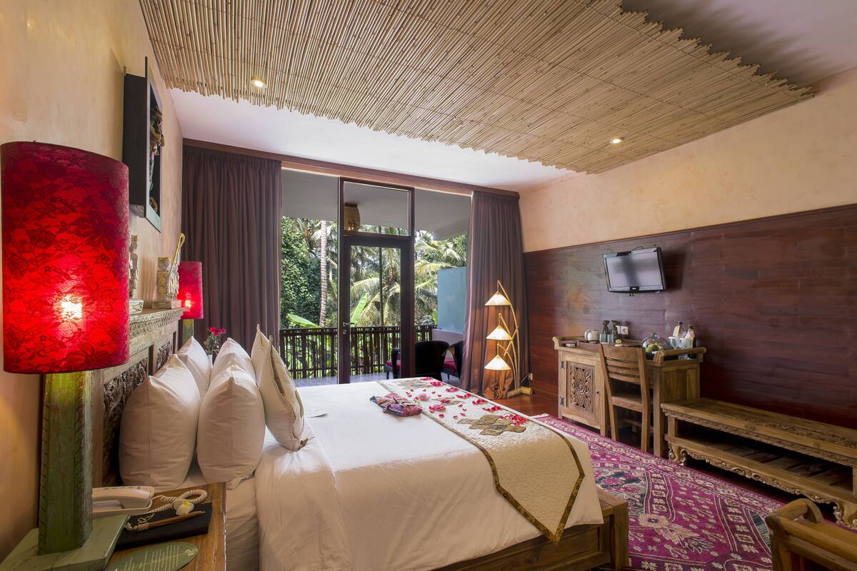 A hotel room with a bed, bedside lamps, a wooden dresser, a television, and a balcony with a view of greenery.