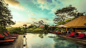 An infinity pool with red lounge chairs and umbrellas, surrounded by lush greenery and a tiled-roof structure.