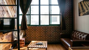 A room with a brick wall, a wooden floor, a brown leather sofa, a wooden coffee table with a remote control, and a window with curtains. - 日木民宿 (Tainan)