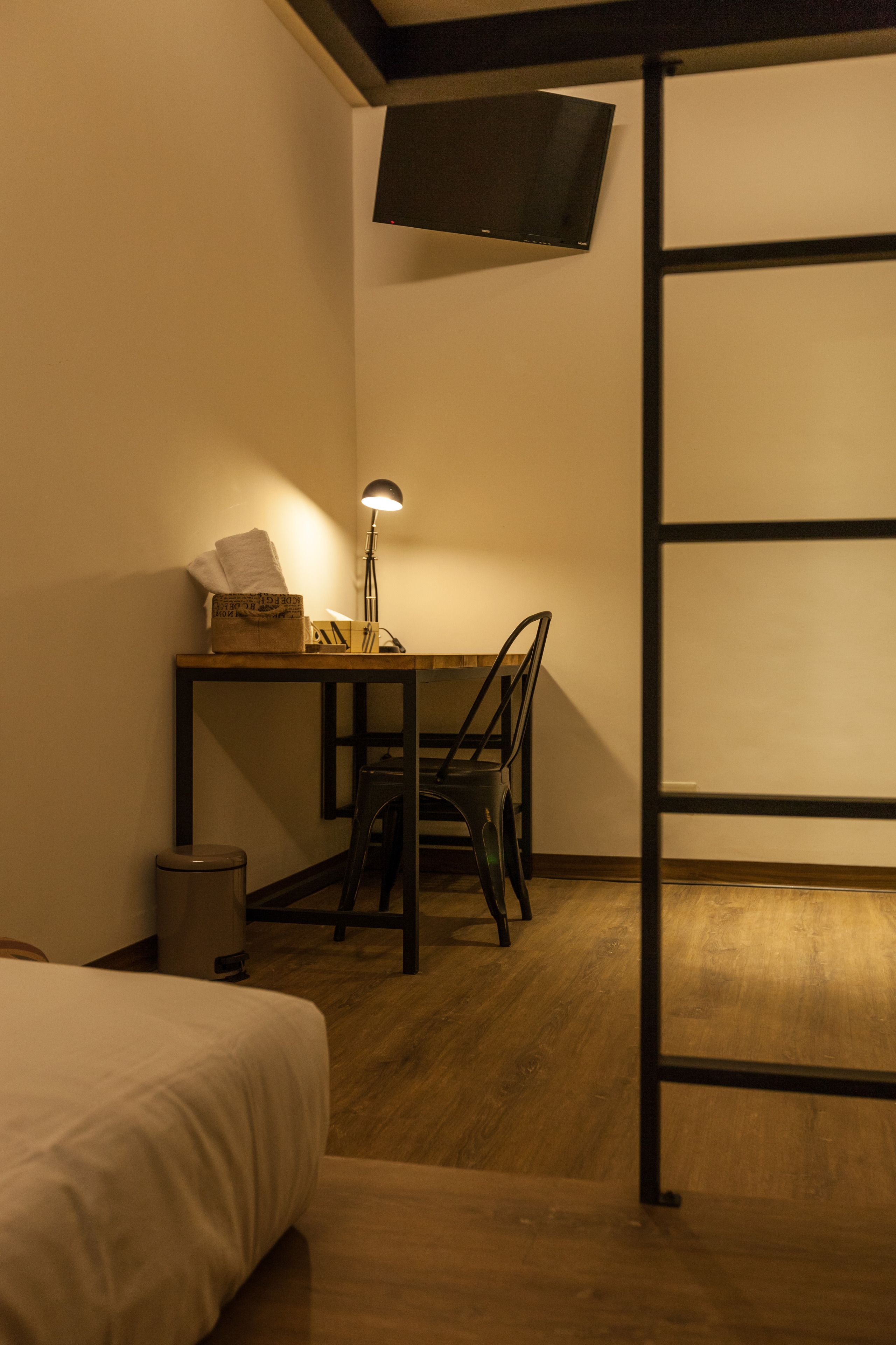A small, dimly lit room with a bed, a desk with a lamp, a chair, and a TV mounted on the wall.