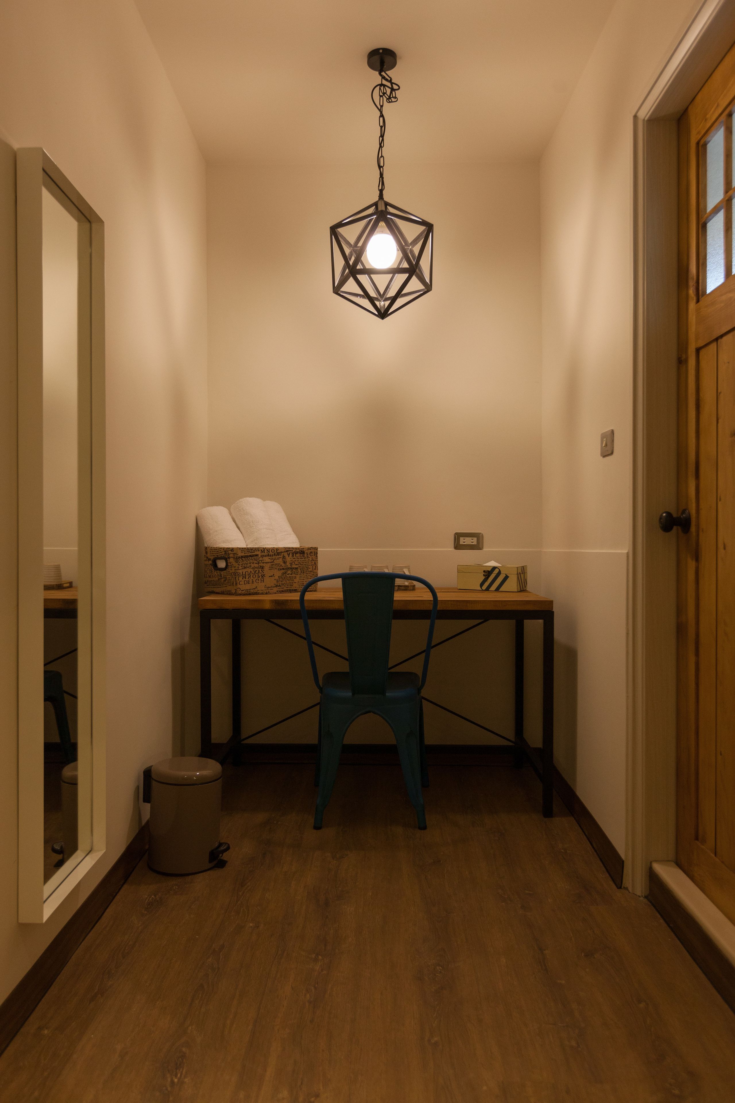 A hallway with a wooden floor, a desk with a chair, a trash can, and a hanging light fixture.
