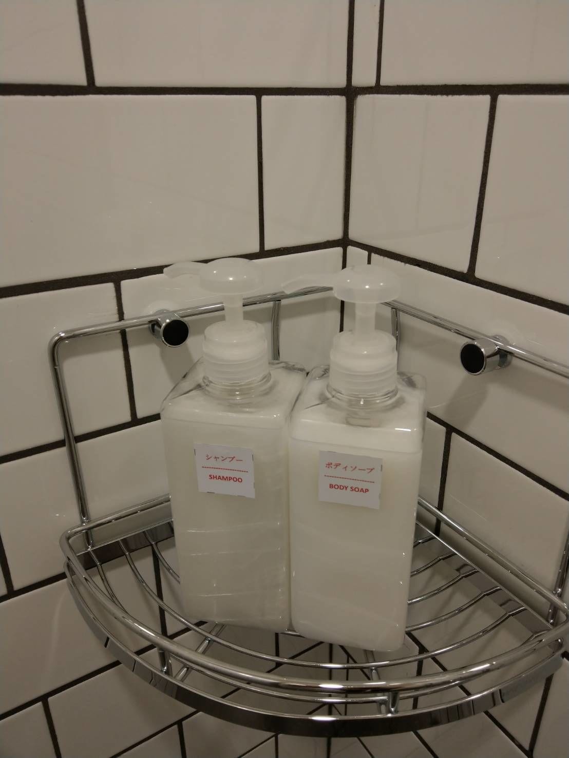 A bathroom with white tiled walls and a metal shelf holding two bottles of shampoo and body soap.