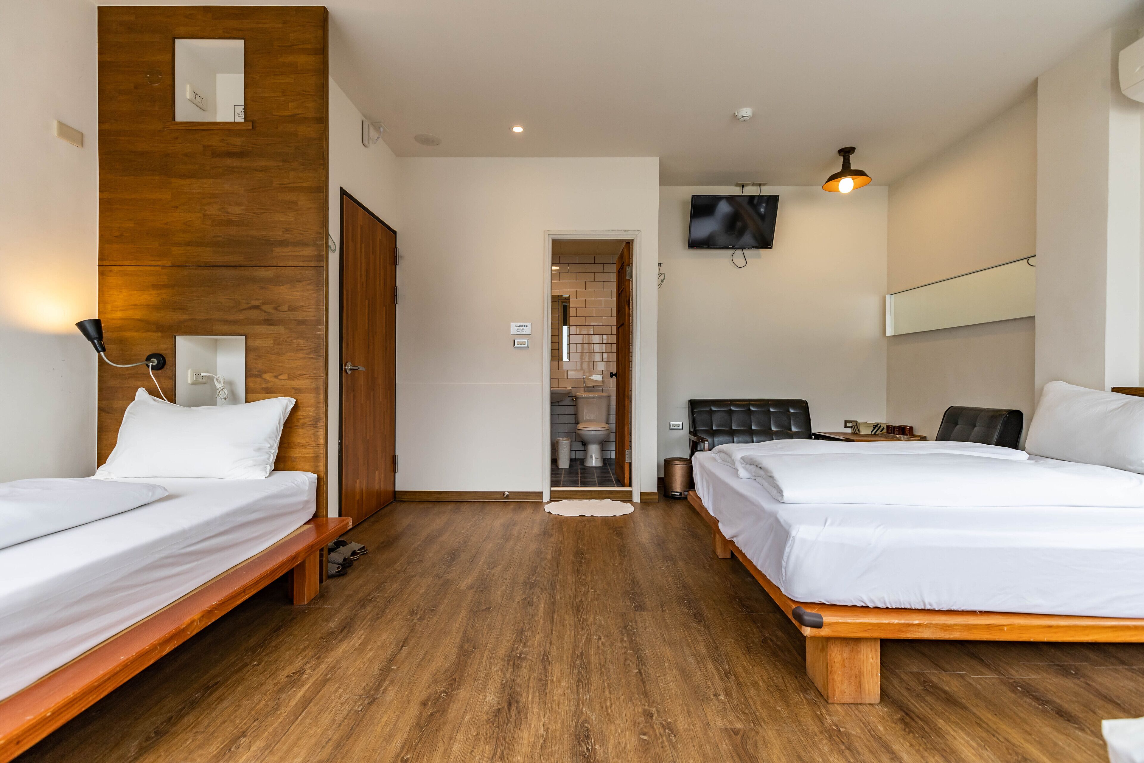 A hotel room with two single beds, a wooden floor, a TV mounted on the wall, and a small desk with a chair.