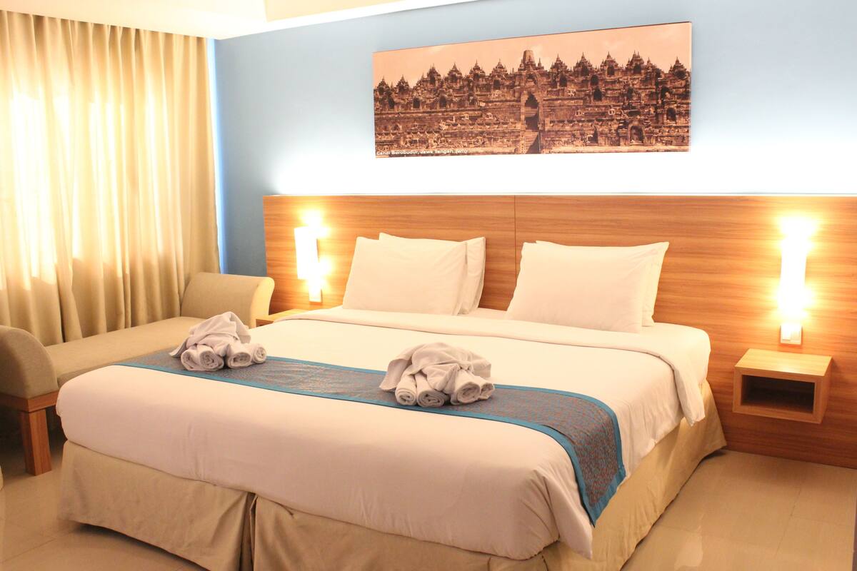 Habitación de hotel con una cama grande, dos mesitas de noche, una silla y un cuadro colgado en la pared.
