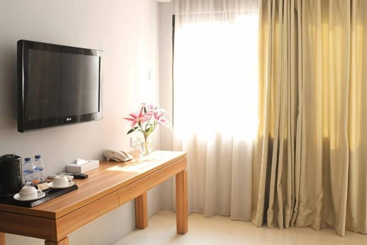 Habitación de hotel con un escritorio de madera, un televisor de pantalla plana, un florero con flores, una ventana con cortinas y una mesita de noche.