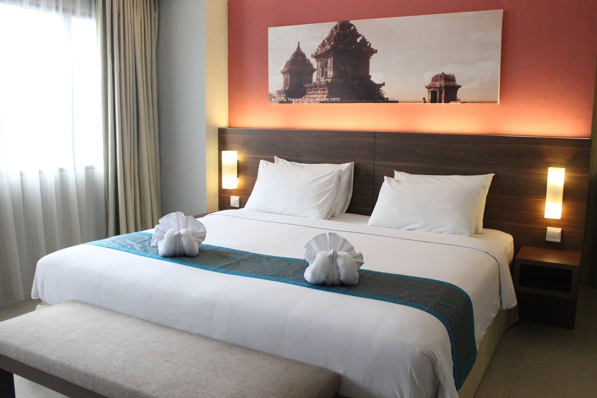 Una habitación de hotel con una cama grande, dos mesitas de noche, un banco y un cuadro colgado en la pared que muestra un templo.