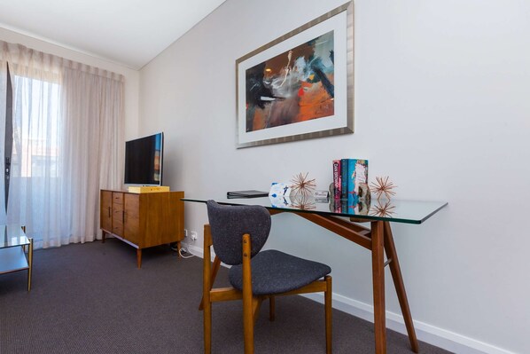 Habitación de hotel moderna con escritorio de vidrio, una silla de madera, televisión y un cuadro abstracto enmarcado en la pared.