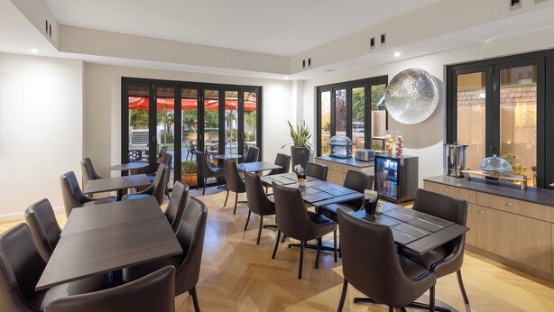 A modern dining area with multiple tables and chairs, a kitchen area with a refrigerator and a coffee machine, and large windows with an outdoor view. - Club Wyndham Perth, Trademark Collection by Wyndham (West Perth)