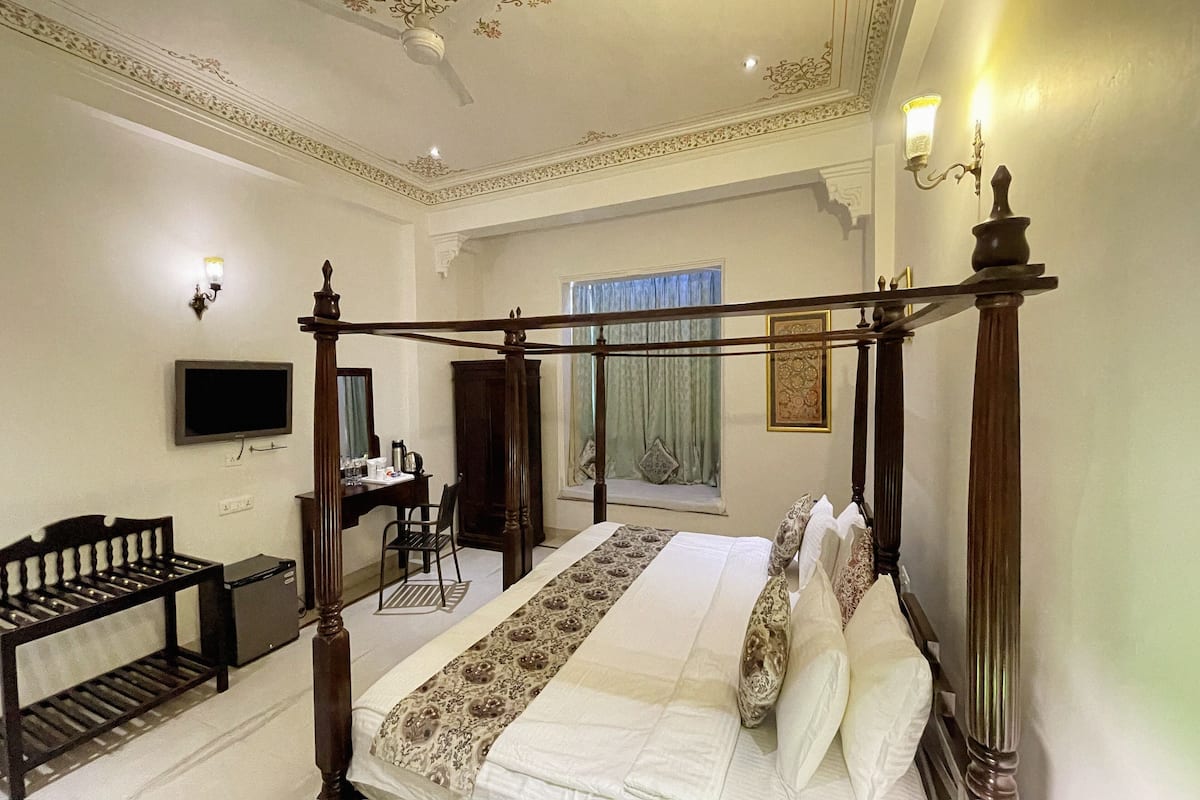 A four-poster bed with a canopy, a television mounted on the wall, a desk with a chair, and a window with curtains.