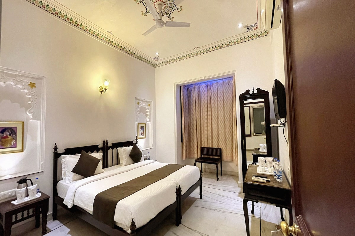 A hotel room with a large bed, a desk, a chair, a mirror, and a ceiling fan.