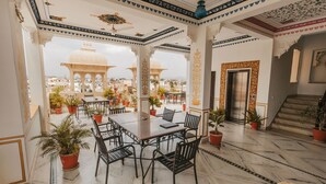 Amplio espacio interior con una mesa grande y sillas, rodeado de plantas en macetas, y vistas a un edificio tradicional.