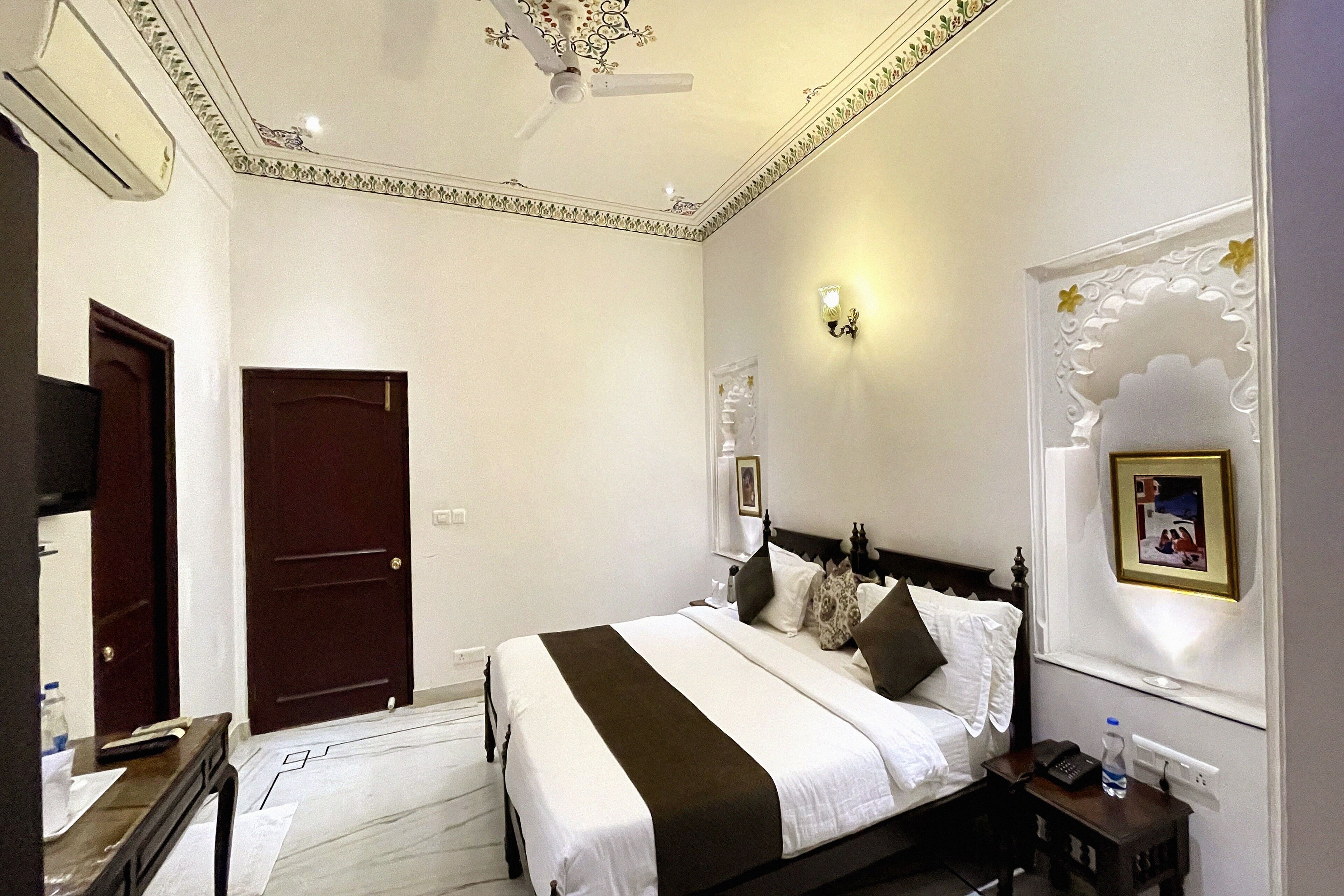 A hotel room with a large bed, a bedside table, a nightstand, a wall-mounted light, and a ceiling fan.