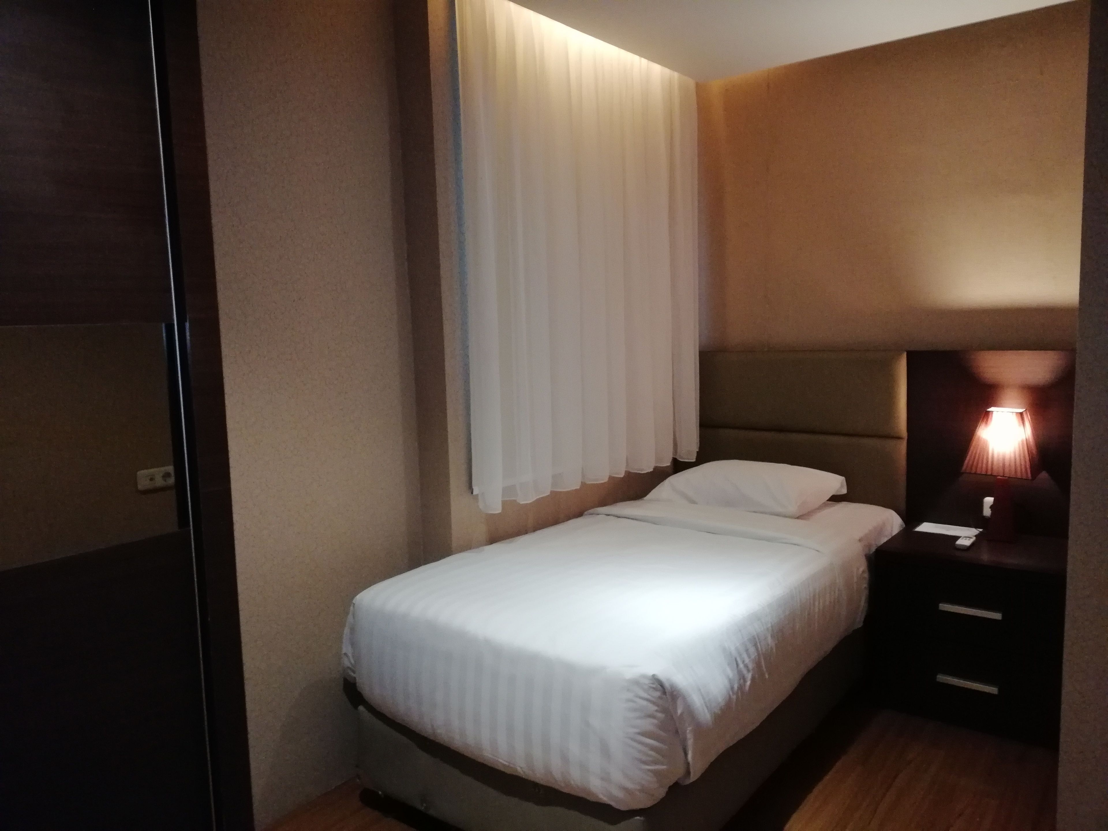 A neatly made bed in a hotel room with a headboard, a bedside table with a lamp, and a wall-mounted curtain.