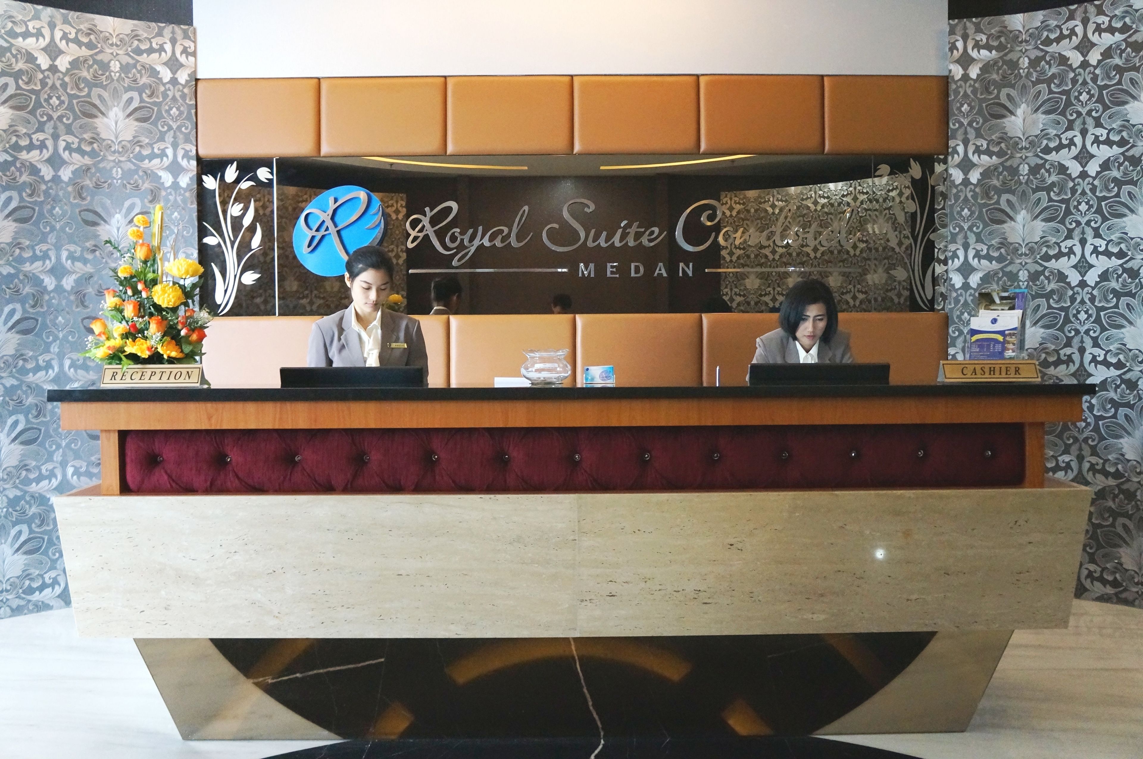 A reception desk at the Royal Suite Co. Medan with a floral arrangement, a cash register, and a sign indicating the cashier area.