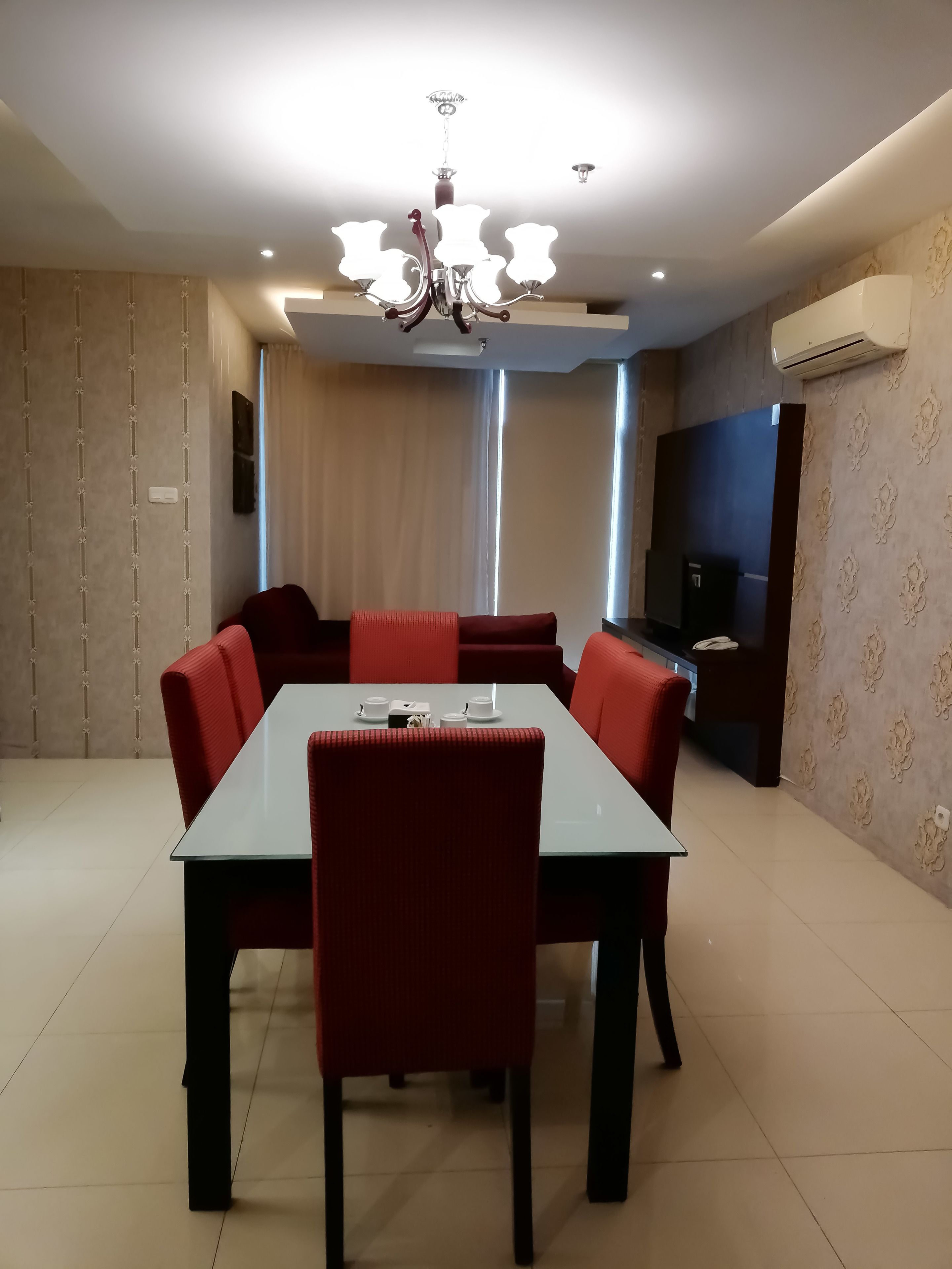 A modern dining area with a rectangular table and red upholstered chairs, a chandelier, and a flat-screen TV mounted on the wall.