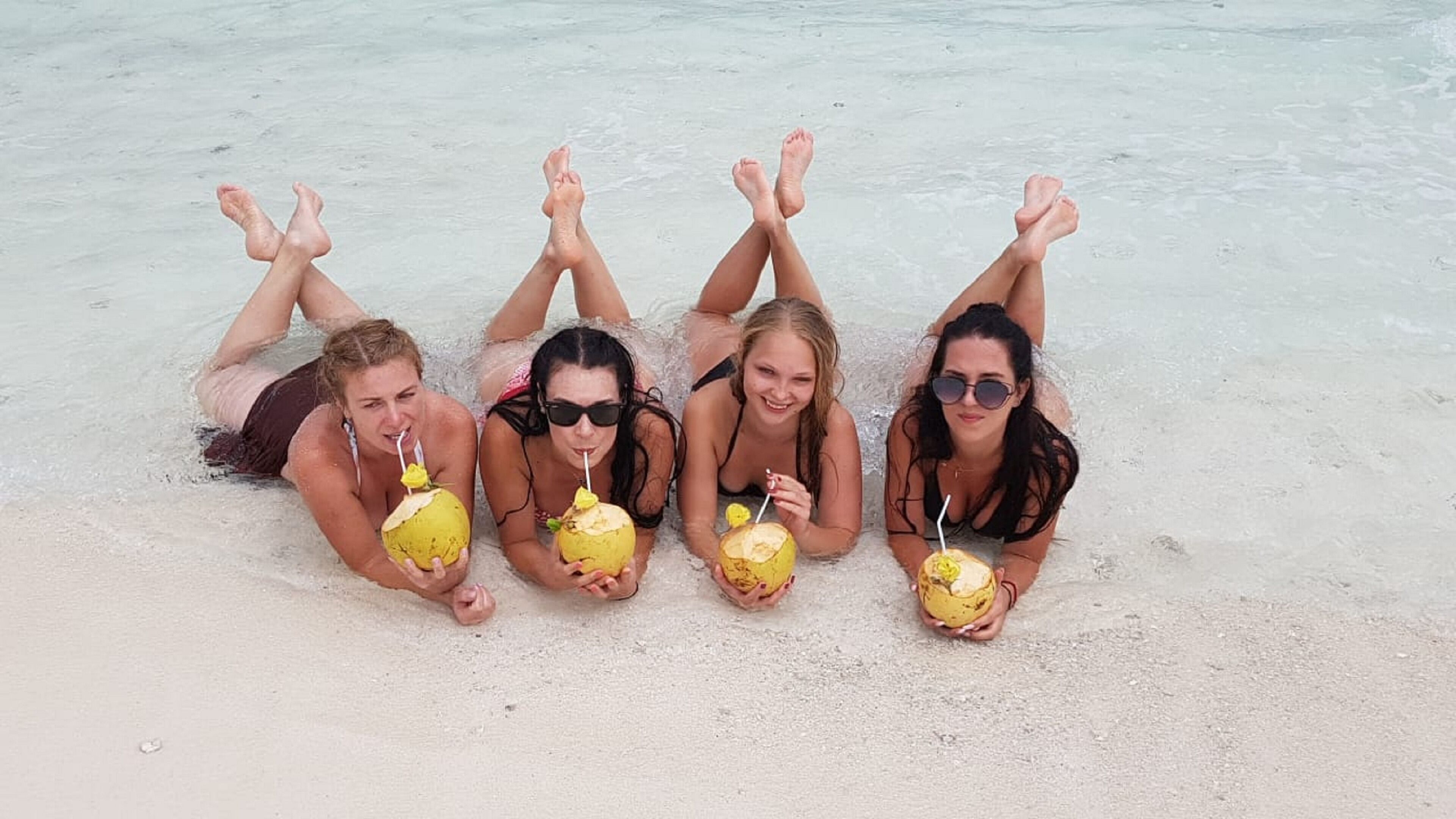 Four people lying on a sandy beach with their feet in the water, each holding a yellow drink.