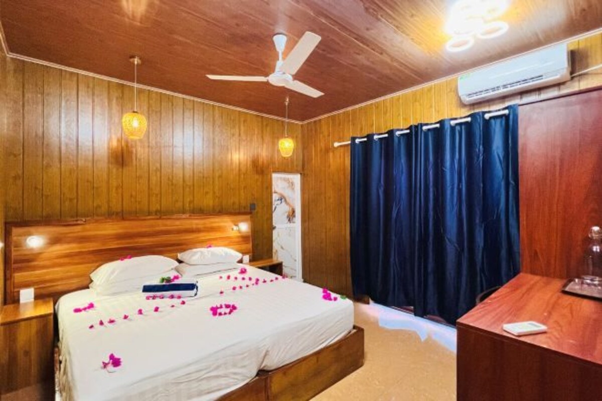 A wooden-paneled bedroom with a bed, a ceiling fan, an air conditioning unit, and a desk with a chair.