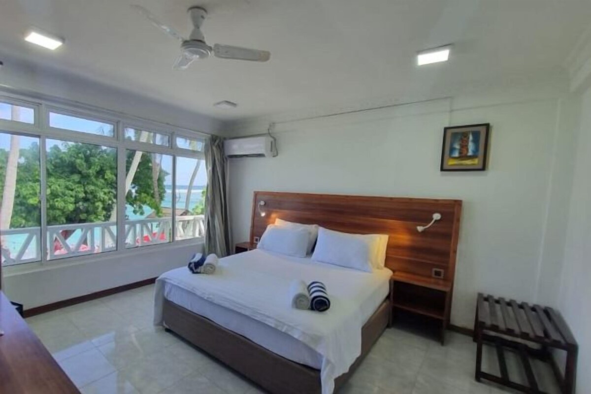 A hotel room with a large bed, a wooden headboard, a nightstand, a bench, and a window with a view of trees.