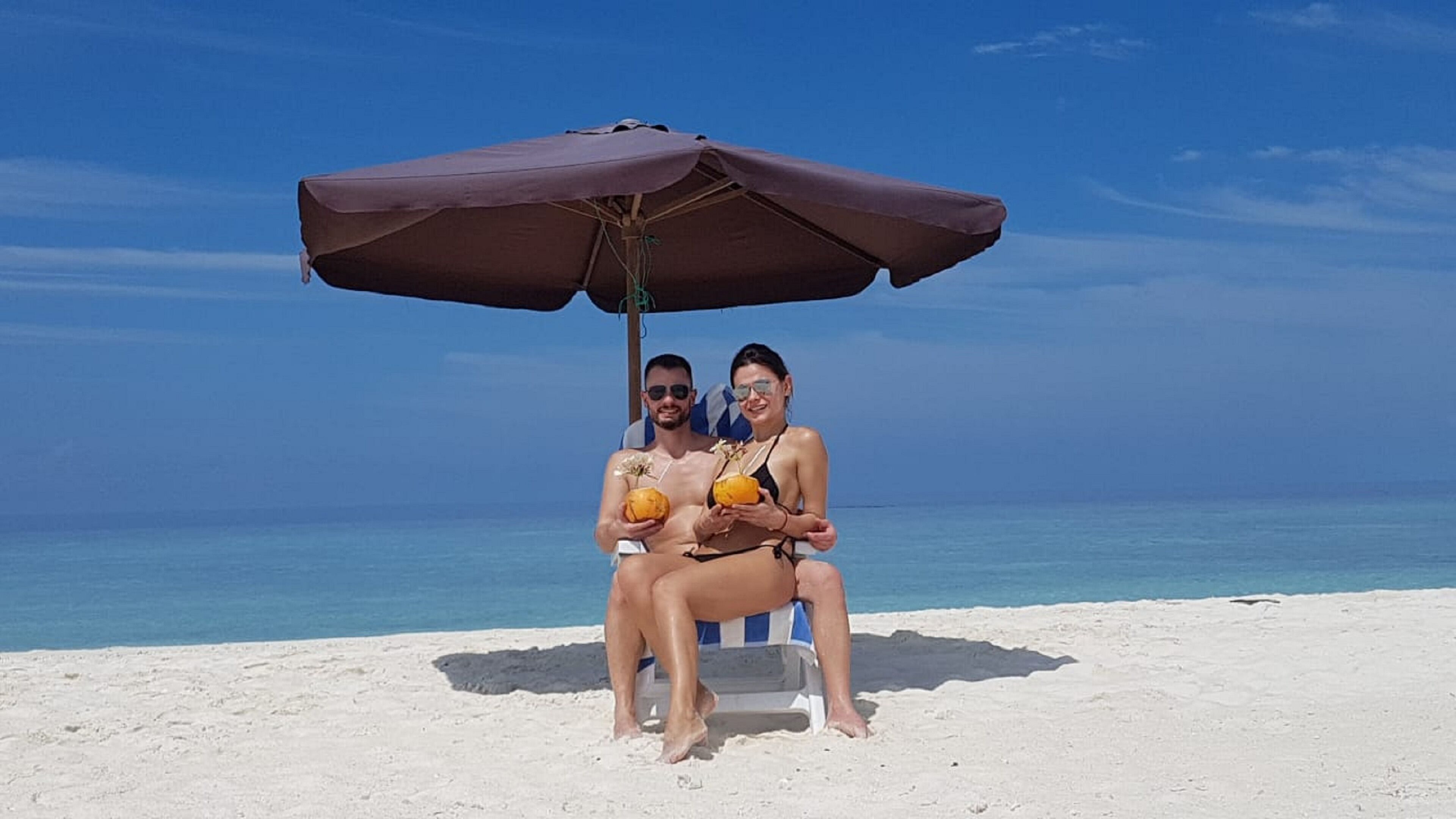 Two people sitting under a beach umbrella on a sandy beach with the ocean in the background.